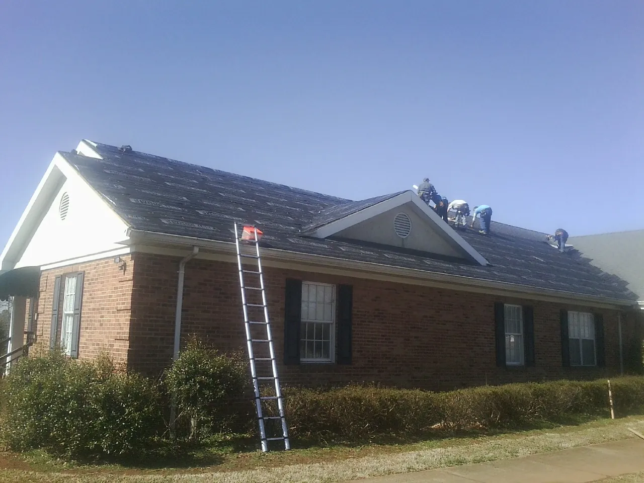 Crew tearing shingles off a brick ranch roof with a ladder set — Emergency Roof Repair work by our crew in East Longmeadow