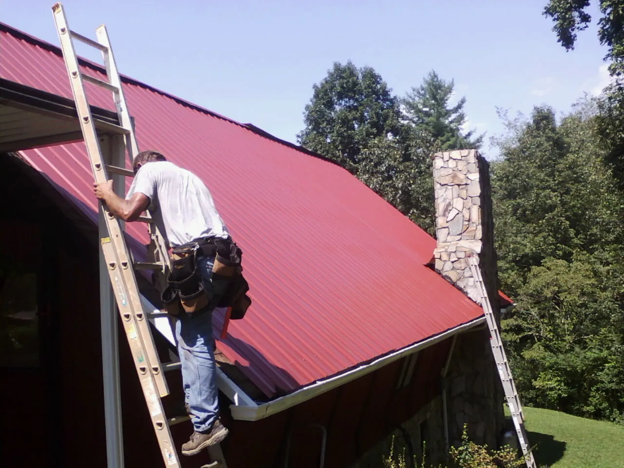 Roofer on a ladder beside a red metal roof and stone chimney — Soffit & Fascia Repair by Roofing Brothers in East Longmeadow